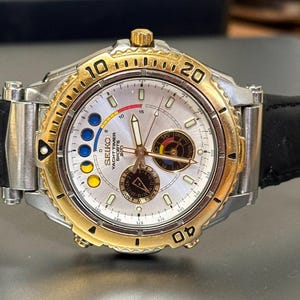 Könnte beinhalten: Eine silber- und goldfarbene Seiko Yacht Timer Sports-Uhr mit schwarzem Lederarmband. Das Zifferblatt ist weiß mit goldenen Akzenten, mit mehreren Hilfszifferblättern und dem Text "SEIKO YACHT TIMER SPORTS 200M".