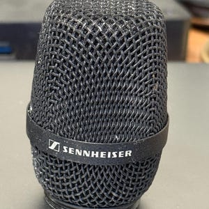 May include: A black Sennheiser microphone head with a woven metal mesh design. The microphone has a black band with the Sennheiser logo. The microphone is on a dark surface.