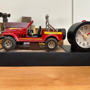 Op de afbeelding: Een rode speelgoedjeep met een zwarte basis en een zwarte wekker. De jeep heeft een gele streep en het nummer 168 aan de zijkant. De wekker heeft een witte wijzerplaat met zwarte cijfers en wijzers.