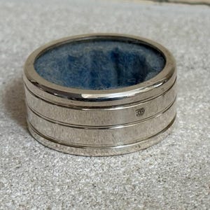 Peut inclure: Une boîte à bagues argentée avec un intérieur bleu. La boîte est de forme cylindrique avec des bandes horizontales. La boîte est posée sur une surface gris clair. Environ 3 cm de diamètre.
