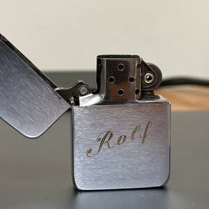 May include: A silver lighter with a brushed finish and the name "Rolf" engraved on the front. The lighter is open and the lid is flipped back.