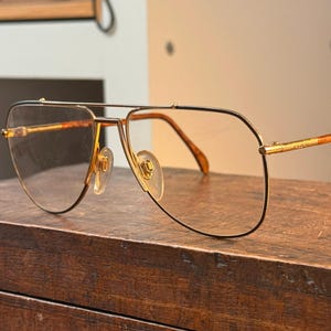 Könnte beinhalten: Goldfarbene Aviator-Brille mit Doppelsteg und klaren Gläsern. Die Fassungen haben einen schwarzen Umriss und braune Bügel. Die Brille liegt auf einer Holzoberfläche. Ein klassisches Brillenaccessoire.