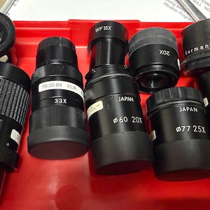 May include: A collection of black microscope lenses and eyepieces, including zoom and fixed magnification models. Some lenses are labeled with "Japan" and "Germany", and magnification strengths like 20x, 25x, 33x, and 40x are visible. The lenses are arranged in a red tray.