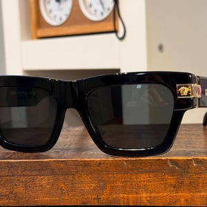 May include: Black sunglasses with a thick, square frame. Gold-colored accents are visible on the temples. The lenses are dark, and the sunglasses are resting on a wooden surface. The overall style is bold and modern.