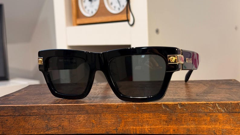 May include: Black sunglasses with a thick, square frame. Gold-colored accents are visible on the temples. The lenses are dark, and the sunglasses are resting on a wooden surface. The overall style is bold and modern.