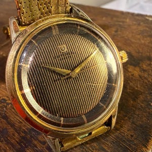 Puede incluir: Reloj de pulsera vintage dorado con esfera marrón a rayas y agujas doradas. El reloj tiene una correa dorada y una caja redonda dorada. El reloj está sobre una superficie de madera.