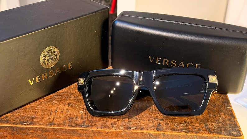 May include: Black sunglasses with a square frame and dark lenses, resting on a wooden surface. The sunglasses are next to a black case and box, both with the Versace logo in gold lettering.