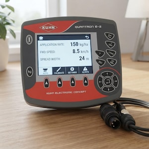 May include: A Kuhn Quantron E-2 electronic control unit with a grey and red housing. The screen displays text including APPLICATION RATE: 150 kg/ha, FWD SPEED: 8.5 km/h, and SPREAD WIDTH: 24 m. It features various buttons, a directional pad, and a black cable.