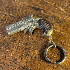Könnte beinhalten: Ein silberfarbener Miniatur-Pistolen-Schlüsselanhänger mit aufwendigen Gravuren. Der Griff ist dunkelbraun, und der Schlüsselanhänger hat einen silbernen Ring. Auf einer strukturierten Holzoberfläche.