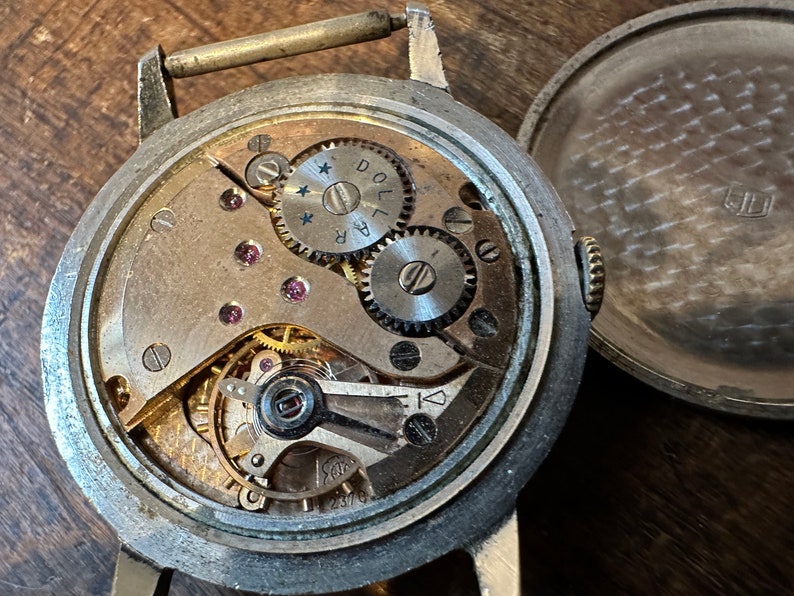Może przedstawiać: Srebrny zegarek kieszonkowy z przezroczystą szybą i widocznym mechanizmem wewnętrznym. Tarcza zegarka zawiera tekst "DOL 14R" i "2370".