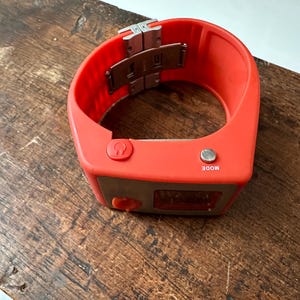 Op de afbeelding: Een feloranje polshorloge met een zilveren gesp en twee knoppen met de tekst "MODE" en een hoefijzer symbool. Het horloge heeft een digitaal display.