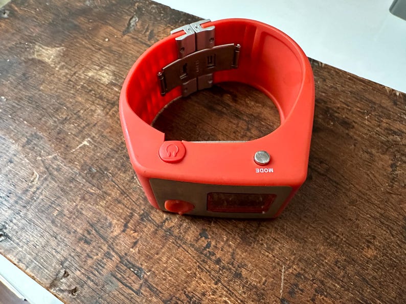 Op de afbeelding: Een feloranje polshorloge met een zilveren gesp en twee knoppen met de tekst "MODE" en een hoefijzer symbool. Het horloge heeft een digitaal display.