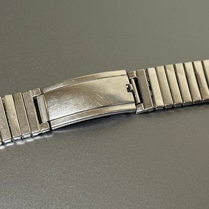 May include: A silver metal watch band with a clasp. The band is made of many small, rectangular links.