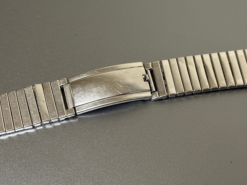 May include: A silver metal watch band with a clasp. The band is made of many small, rectangular links.