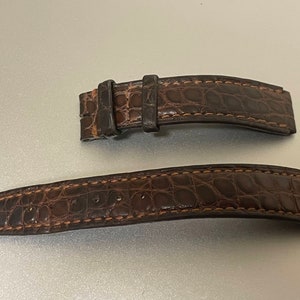 May include: Two-piece brown watch strap made of textured leather, possibly alligator or crocodile. The strap is dark brown with lighter brown stitching. The buckle is visible on one end.