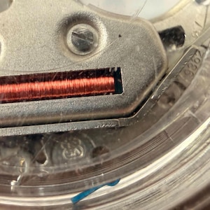 Puede incluir: Primer plano de un mecanismo de reloj, que muestra detalles intrincados. Una bobina de color cobre es visible dentro de un componente met&aacute;lico, rodeada por una carcasa transparente. La imagen destaca la precisi&oacute;n y complejidad del funcionamiento interno del reloj.