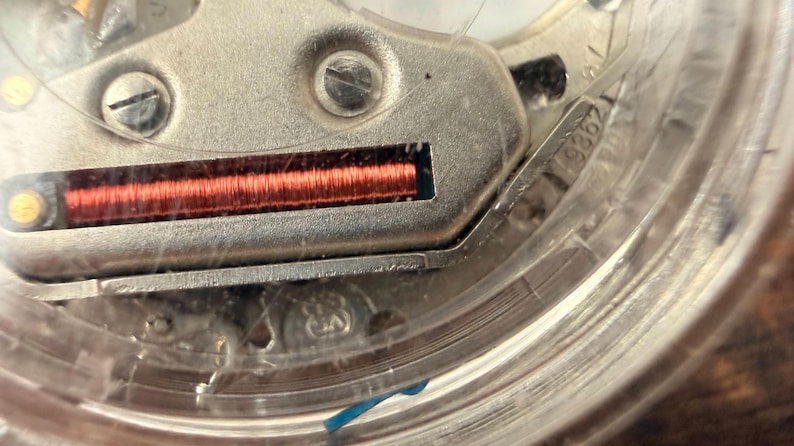 Puede incluir: Primer plano de un mecanismo de reloj, que muestra detalles intrincados. Una bobina de color cobre es visible dentro de un componente met&aacute;lico, rodeada por una carcasa transparente. La imagen destaca la precisi&oacute;n y complejidad del funcionamiento interno del reloj.