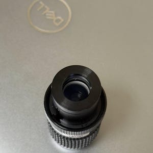 Puede incluir: Un objetivo de c&aacute;mara negro con un anillo de ajuste plateado y acanalado. La lente es cil&iacute;ndrica con una abertura central. La lente est&aacute; sobre una superficie gris de ordenador port&aacute;til con el logotipo de Dell visible.
