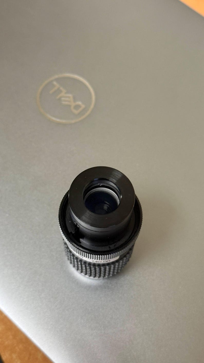Puede incluir: Un objetivo de c&aacute;mara negro con un anillo de ajuste plateado y acanalado. La lente es cil&iacute;ndrica con una abertura central. La lente est&aacute; sobre una superficie gris de ordenador port&aacute;til con el logotipo de Dell visible.