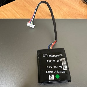 May include: A black battery with a white label that reads "Microsemi ASCM-35F 5.4V 35F Patent # US 9,525,298". The battery has a green dot on the label and a white connector with wires.