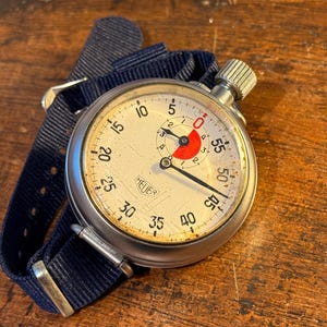 Op de afbeelding: Een vintage zilveren stopwatch met een witte wijzerplaat en zwarte markeringen. De stopwatch heeft een zwarte nylon band en een rode accent op de wijzerplaat. De wijzerplaat heeft "HEUER" en heeft nummers van 5 tot 60.