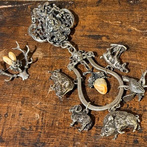 Peut inclure: Un pendentif argenté orné avec divers breloques d'animaux, dont un bélier, un sanglier et un oiseau. Le pendentif comprend également une pierre centrale et une broche en forme de bois de cerf avec une abeille. Le bijou est présenté sur une surface en bois.