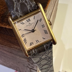 May include: A rectangular gold-toned wristwatch with a light-coloured face and black Roman numeral hour markers. The watch features the brand name "Baume & Mercier Geneve" and a grey, textured leather strap. The watch is Swiss made.