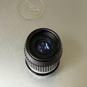 Puede incluir: Un objetivo de c&aacute;mara negro con un anillo de ajuste plateado y acanalado. El objetivo est&aacute; sobre una superficie plateada de ordenador port&aacute;til con un logotipo de Dell.