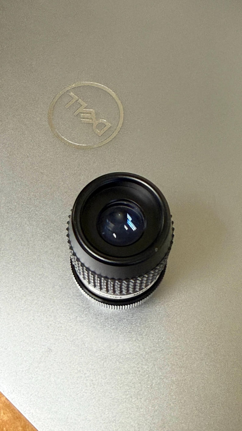 Puede incluir: Un objetivo de c&aacute;mara negro con un anillo de ajuste plateado y acanalado. El objetivo est&aacute; sobre una superficie plateada de ordenador port&aacute;til con un logotipo de Dell.