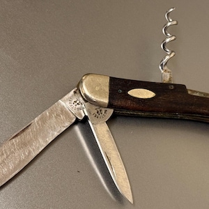 Puede incluir: Una navaja de bolsillo vintage con sacacorchos y dos hojas. La navaja tiene un mango de madera marrón oscuro con un acento plateado. Las hojas están abiertas, mostrando desgaste. El sacacorchos es plateado.