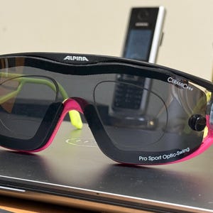 Könnte beinhalten: Schwarze und pinkfarbene Sportsonnenbrille mit dem Markennamen "ALPINA" oben. Die Sonnenbrille hat ein umlaufendes Design mit dunklen Gläsern und neongrünen und gelben Akzenten. Der Text "Pro Sport Optic-Swing" ist sichtbar.