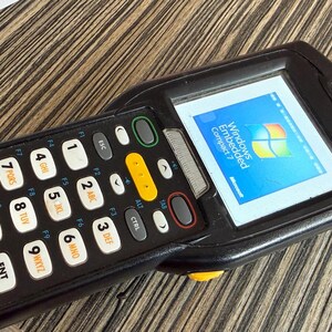 Puede incluir: Un ordenador de mano negro con teclado y pantalla que muestra el logotipo de Windows Embedded Compact 7. El dispositivo tiene el logotipo "symbol" y varias teclas de función. El teclado tiene teclas blancas con números y letras.
