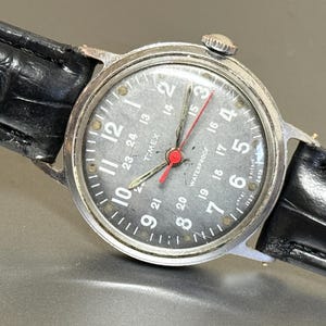 Könnte beinhalten: Eine Vintage-Armbanduhr von Timex mit silberfarbenem Gehäuse und dunkelgrauem Zifferblatt. Die Uhr hat weiße Ziffern, einen roten Sekundenzeiger und ein schwarzes Lederarmband. Das Zifferblatt zeigt das Wort "TIMEX" und "WATERPROOF".