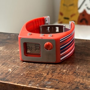 Op de afbeelding: Een rood-blauw gestreept digitaal polshorloge met een zilveren wijzerplaat en een grote knop aan de zijkant. De wijzerplaat van het horloge geeft de tijd 22:00 aan.