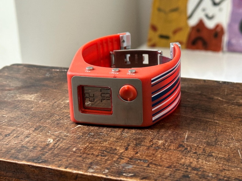 Op de afbeelding: Een rood-blauw gestreept digitaal polshorloge met een zilveren wijzerplaat en een grote knop aan de zijkant. De wijzerplaat van het horloge geeft de tijd 22:00 aan.