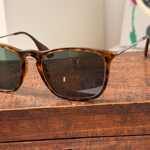 May include: Sunglasses with a tortoiseshell frame and dark lenses are displayed on a wooden surface. The sunglasses have a classic square shape with a brown and tan pattern. The temples are dark brown and the arms are silver.