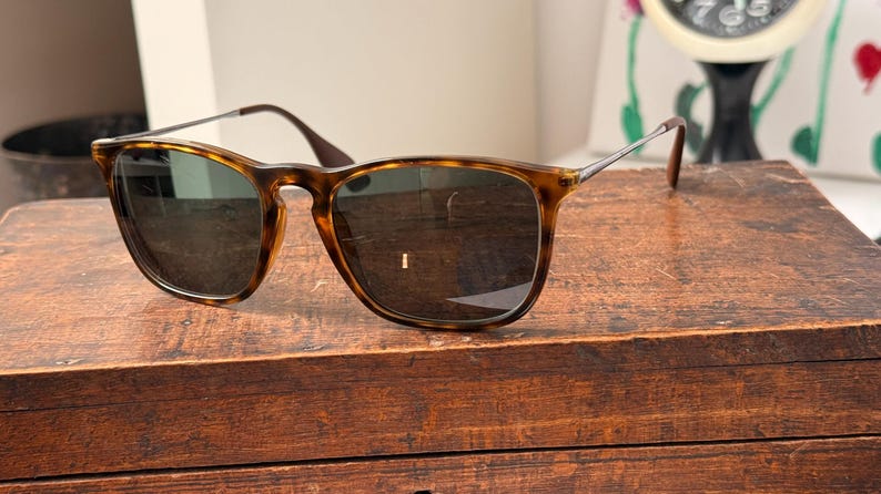 May include: Sunglasses with a tortoiseshell frame and dark lenses are displayed on a wooden surface. The sunglasses have a classic square shape with a brown and tan pattern. The temples are dark brown and the arms are silver.
