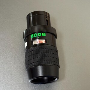 Puede incluir: Un objetivo zoom negro con texto verde "ZOOM" y "60X". El objetivo tiene un agarre estriado y un cuerpo cil&iacute;ndrico liso. El objetivo est&aacute; dise&ntilde;ado para su uso con una c&aacute;mara.
