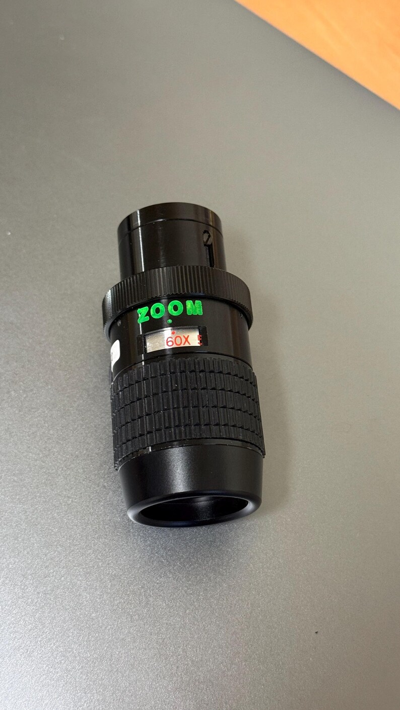 Puede incluir: Un objetivo zoom negro con texto verde "ZOOM" y "60X". El objetivo tiene un agarre estriado y un cuerpo cil&iacute;ndrico liso. El objetivo est&aacute; dise&ntilde;ado para su uso con una c&aacute;mara.