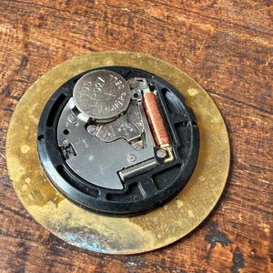 May include: Close-up of the back of a round, gold-colored clock face. The clock face has a black circular rim with a battery and a small metal spring inside. The battery is labeled "392".