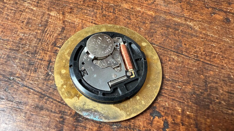 May include: Close-up of the back of a round, gold-colored clock face. The clock face has a black circular rim with a battery and a small metal spring inside. The battery is labeled "392".