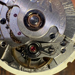 May include: Close-up of a silver watch movement with a red jewel and the text "2834-2" visible. The movement is intricate and detailed, showcasing the inner workings of a mechanical watch.
