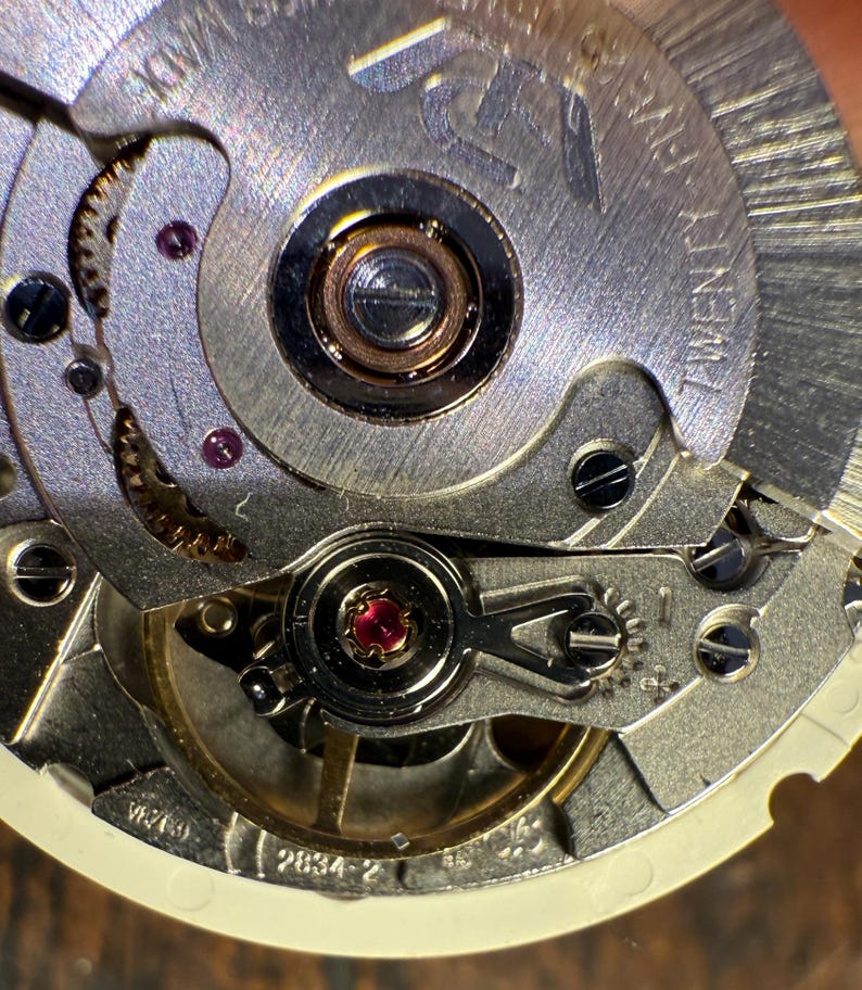 May include: Close-up of a silver watch movement with a red jewel and the text "2834-2" visible. The movement is intricate and detailed, showcasing the inner workings of a mechanical watch.