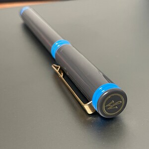 May include: A black pen with blue accents and a gold clip. The pen has a gold cap with a logo that says "W" in a circle.