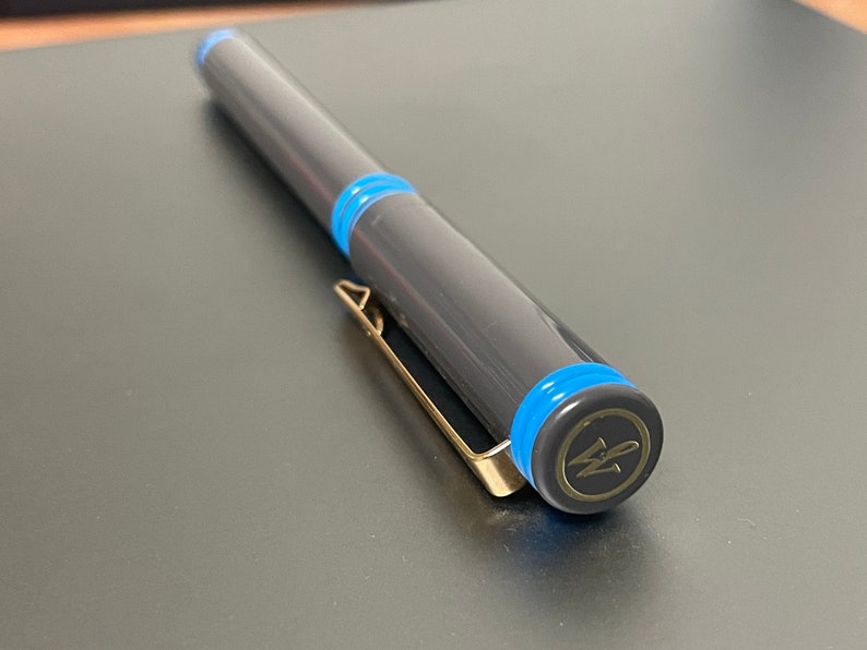 May include: A black pen with blue accents and a gold clip. The pen has a gold cap with a logo that says "W" in a circle.