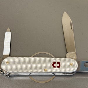 Könnte beinhalten: Ein silberfarbenes Multifunktions-Taschenmesser mit strukturiertem Alox-Griff und einem rot-weißen Schweizer Kreuz-Emblem. Das Messer hat mehrere ausgeklappte Edelstahlwerkzeuge, darunter eine Hauptklinge, eine kleine Klinge und einen Flaschenöffner.