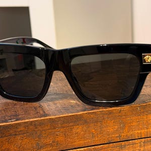 May include: Black square-framed sunglasses with dark lenses. The sunglasses have gold-colored accents on the temples and a gold emblem on the side. The sunglasses are displayed on a wooden surface.