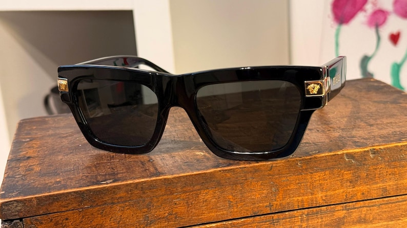 May include: Black square-framed sunglasses with dark lenses. The sunglasses have gold-colored accents on the temples and a gold emblem on the side. The sunglasses are displayed on a wooden surface.