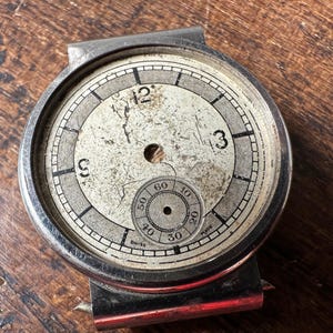 Peut inclure: Un cadran de montre argenté avec des marques noires et un petit sous-cadran circulaire. Le cadran de la montre est fissuré et montre des signes d'usure. Le texte "SWISS MADE" est visible sur le sous-cadran.