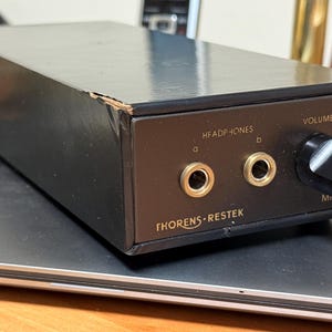 May include: Black Thorens-Restek headphone amplifier with a volume knob and two headphone jacks. The front panel has the text "HEADPHONES" and "VOLUME". The amplifier is on a dark surface.